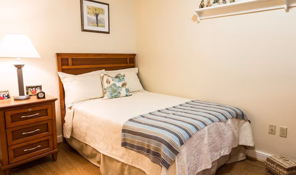 A cozy bedroom with a wooden bed frame, a neatly made bed with white bedding and a striped throw blanket. There is a wooden nightstand with a lamp, two framed photos, and a small clock. A framed picture of a tree hangs on the wall above the bed, and a shelf with decorative figurines is mounted on the adjacent wall.