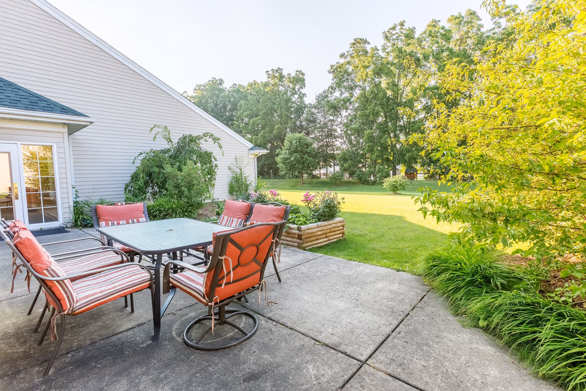 Outdoor patio area with a glass-top table surrounded by six cushioned chairs with red and striped upholstery. The patio is adjacent to a light gray building with a door and windows. There is a small raised garden bed with flowers and green grass extending into a yard with trees in the background.