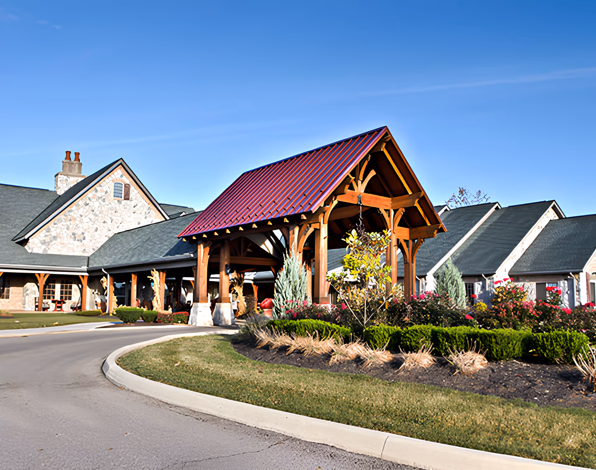 Exterior view of Heritage Senior Living of Marysville showing a curved driveway leading to a covered entrance with wooden beams and a red roof. The building has stone and brick walls with multiple gabled roofs. There are landscaped bushes and flowers around the entrance area under a clear blue sky.