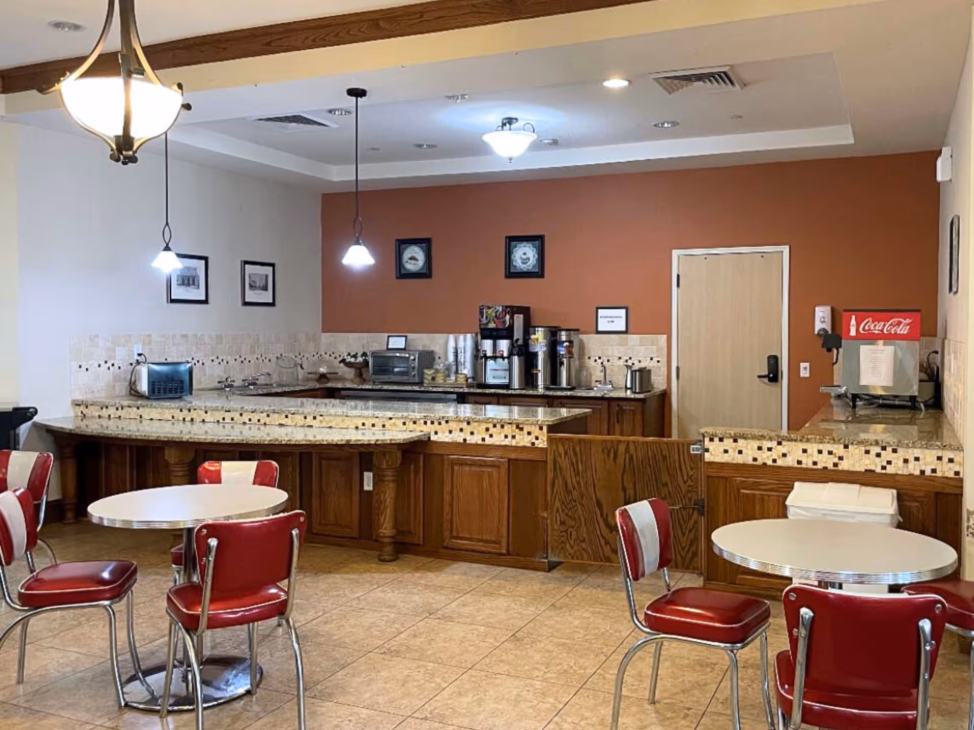 A small dining area with round tables and red cushioned chairs. The background features a counter with a coffee station, toaster oven, and a Coca-Cola beverage dispenser. The walls are painted white and a warm brown color, with framed pictures hanging. The floor is tiled, and there are pendant lights and a ceiling light fixture.