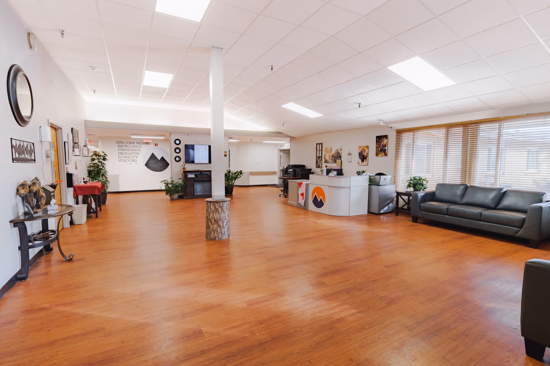 Spacious reception lobby in a senior care facility with wood floors, a reception desk, couch seating, and large windows with blinds.