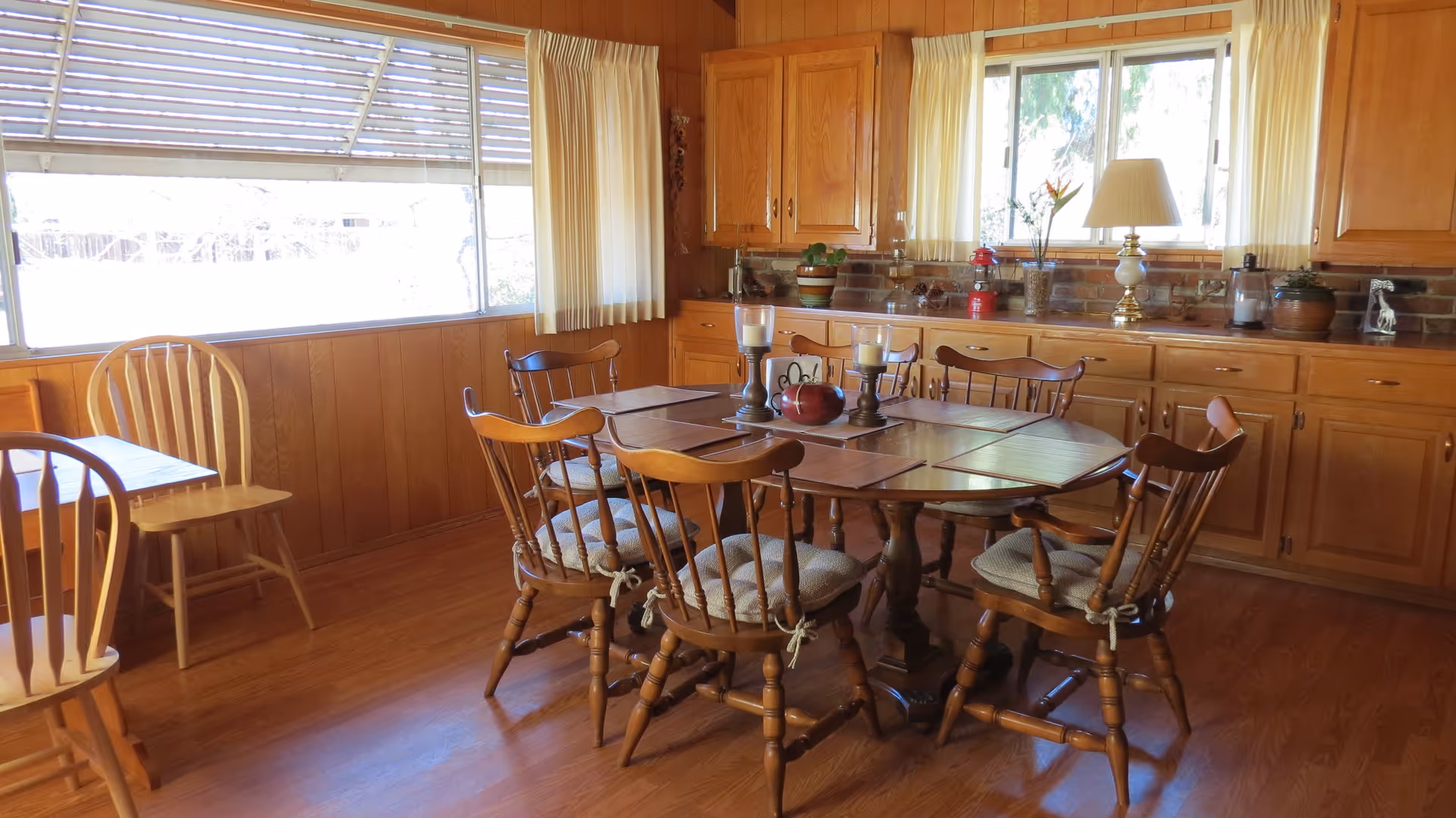 A cozy dining room with a wooden table surrounded by six wooden chairs with cushions. The room has wood-paneled walls, large windows with cream-colored curtains, and built-in wooden cabinets along one wall. The table is decorated with placemats, two candle holders, and a small decorative pumpkin. A lamp and various small plants and decorations are placed on the cabinet countertop.