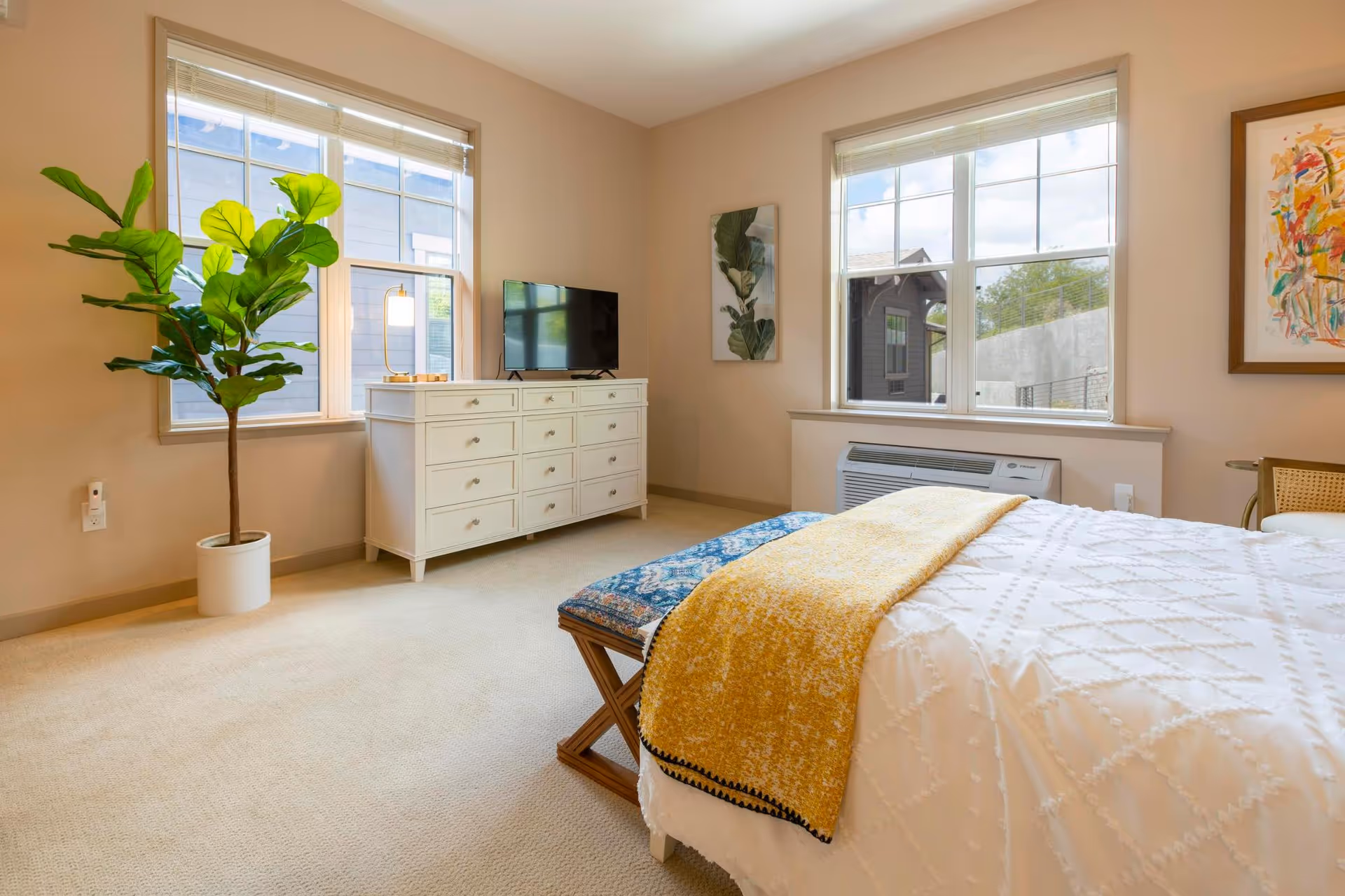 A bright bedroom with two large windows, a white dresser with a TV on top, a potted plant, a bench with a blue cushion at the foot of the bed, and a bed covered with a white textured blanket and a yellow throw. The walls are beige, and there are two pieces of wall art visible.
