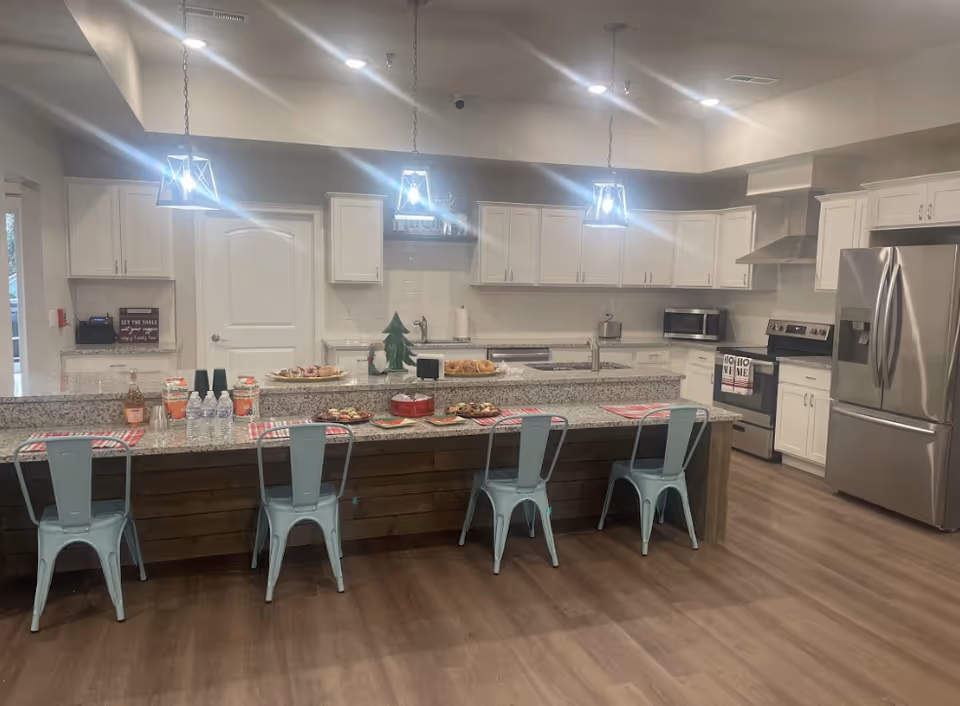 Open communal kitchen with a long granite island, light-blue metal bar stools, white cabinets, stainless appliances and pendant lights.