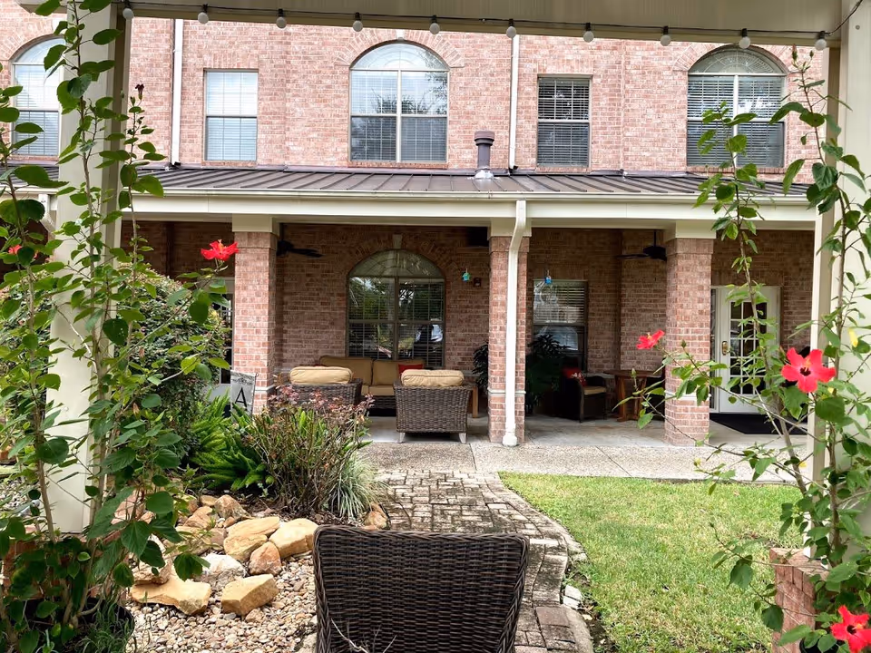 Outdoor patio area with wicker chairs and a sofa with beige cushions under a covered porch attached to a brick building. There are green plants and red flowers in the foreground, a stone and pebble garden bed, and a grassy lawn area.