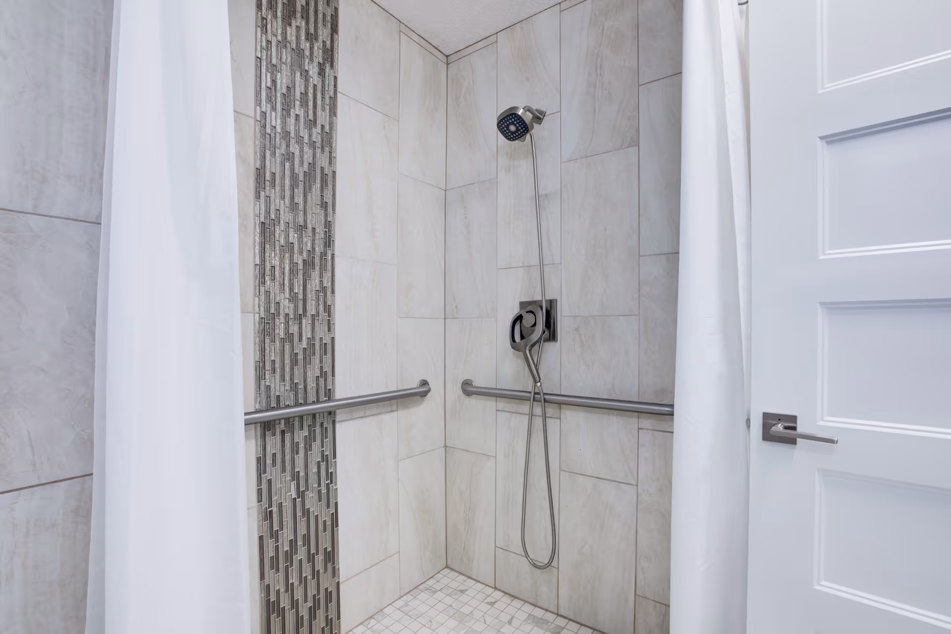 Accessible tiled shower stall with grab bars, a handheld showerhead, mosaic accent tile and white curtains.