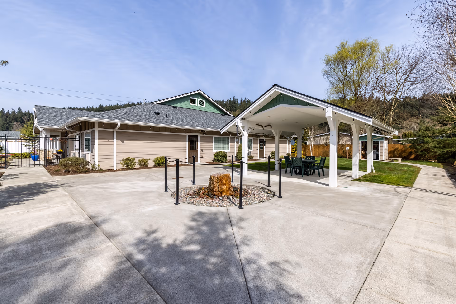 Outdoor courtyard area of a senior living facility with a covered seating area containing a table and chairs, surrounded by concrete walkways, grass, and trees. The building has beige siding with green accents and a gray roof under a clear blue sky.