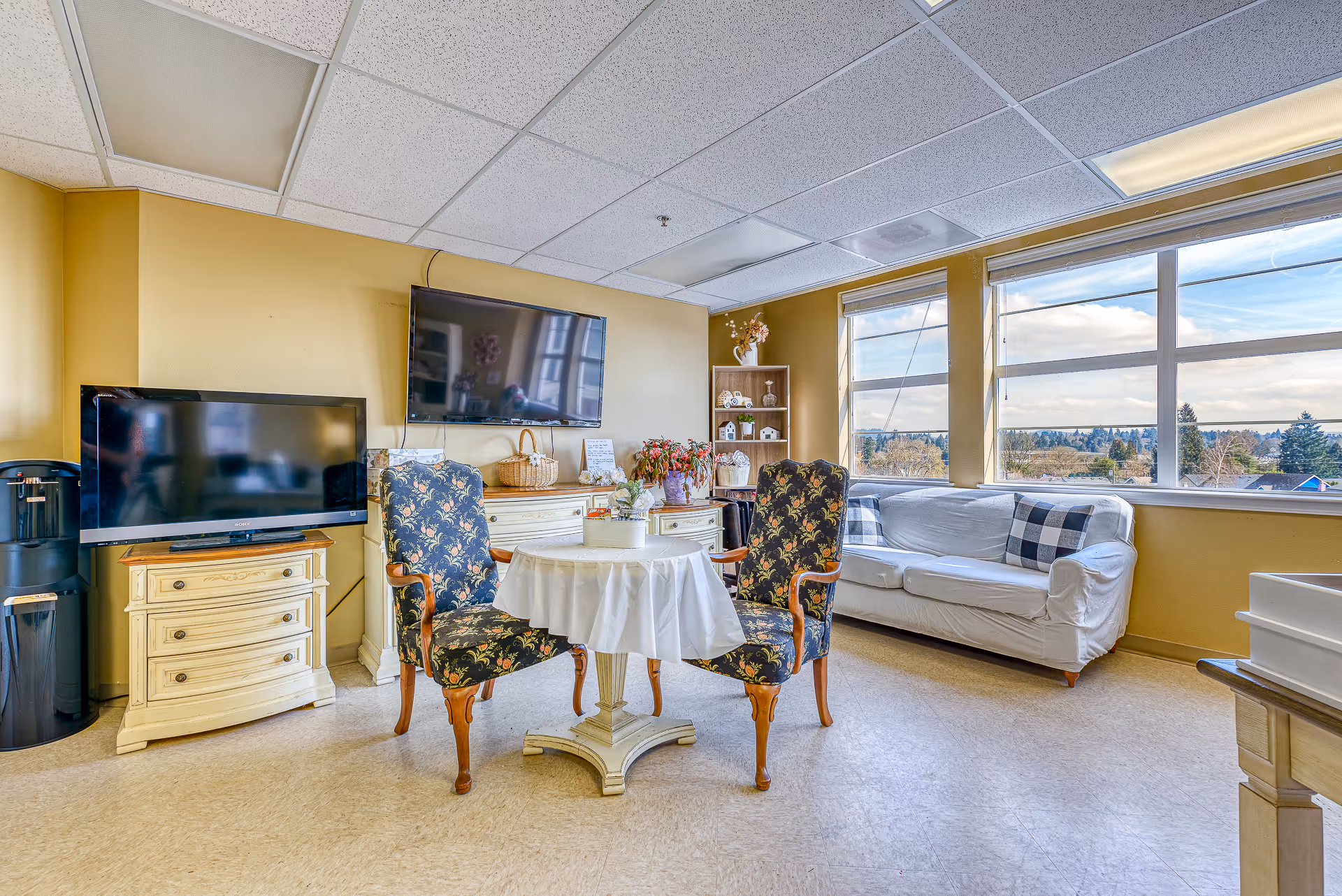 A cozy senior living common area with two floral upholstered armchairs around a small round table covered with a white tablecloth. Behind the chairs is a white couch with plaid pillows next to large windows showing a scenic outdoor view. Two flat-screen TVs are mounted on the walls above cream-colored cabinets. The room has a beige floor and yellow walls.