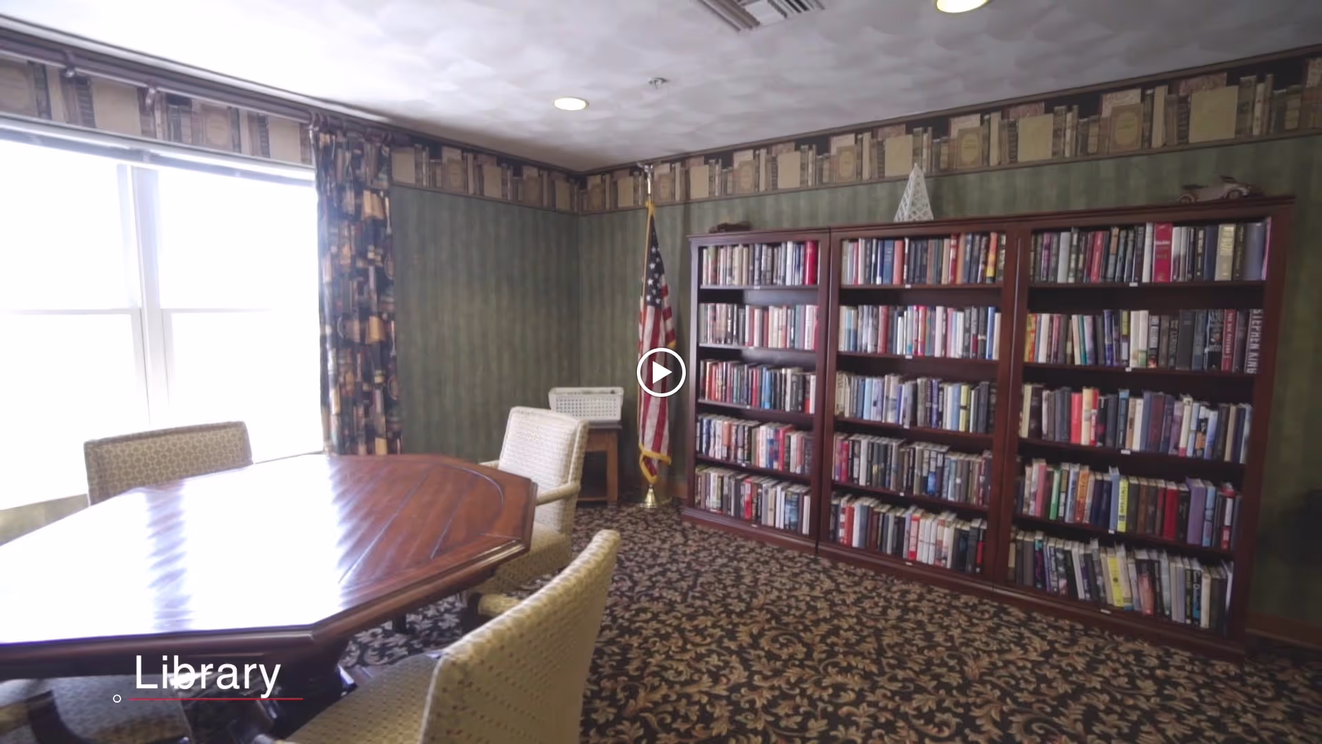 A cozy library room with a wooden table surrounded by cushioned chairs, a large bookshelf filled with books, an American flag in the corner, patterned carpet, and a window with patterned curtains allowing natural light to enter.