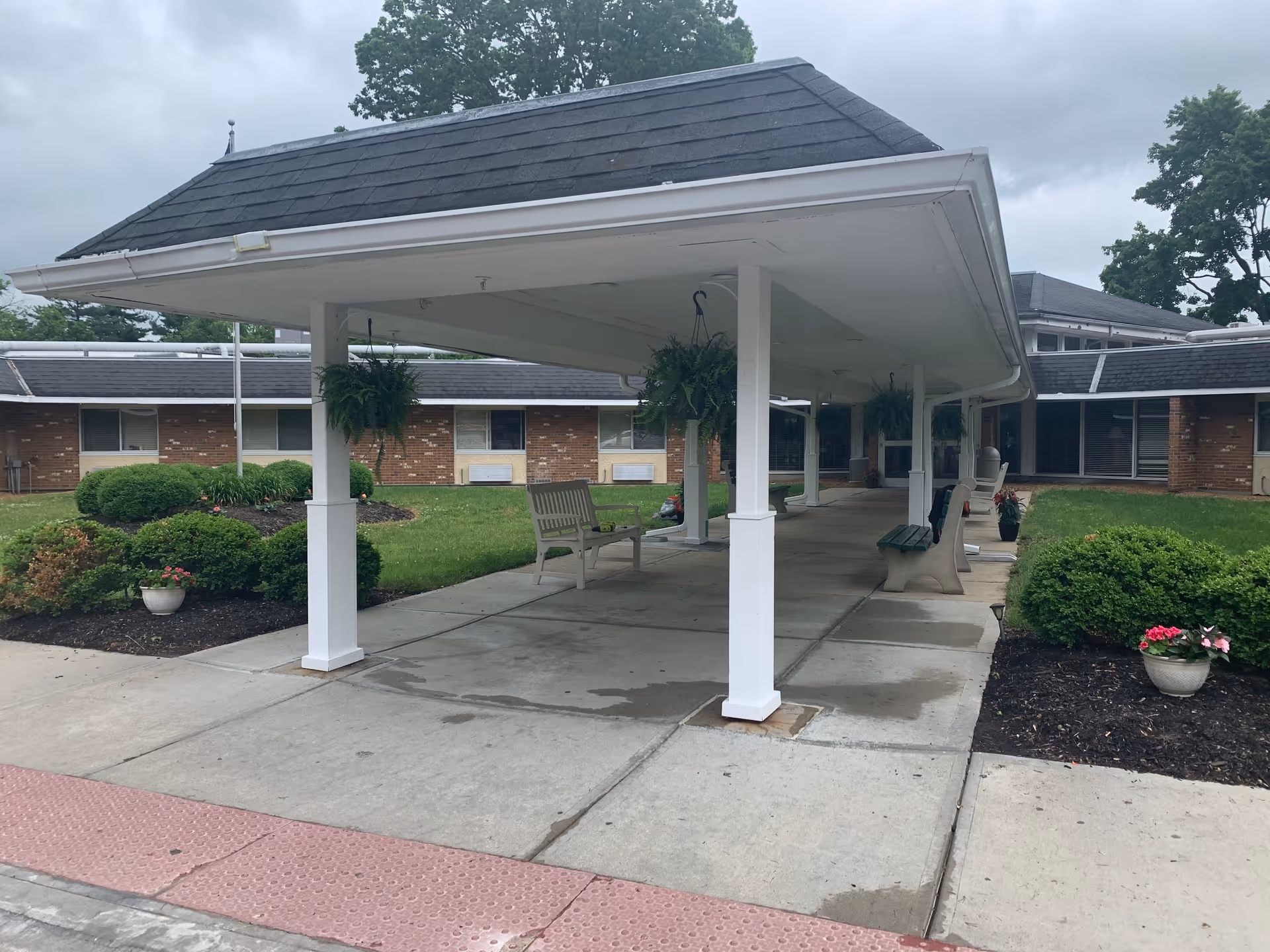 Covered outdoor walkway with white pillars and hanging plants, benches along the sides, surrounded by green bushes and a brick building in the background under a cloudy sky.