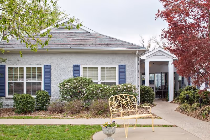 Front exterior of a single-story building with blue-shuttered windows, a walkway, landscaping, and a yellow metal bench.