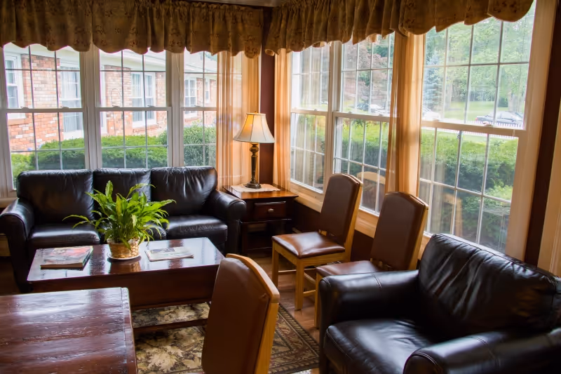 A cozy living room area with large windows letting in natural light. The room features two black leather sofas, two wooden chairs with brown cushions, a wooden coffee table with a potted plant and magazines on it, a side table with a lamp, and beige curtains with a valance. Outside the windows, greenery and a brick building are visible.