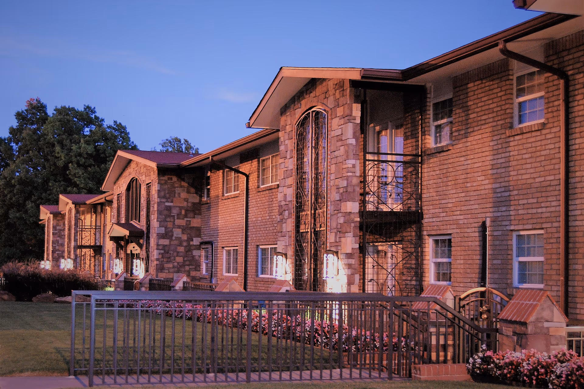Two-story brick retirement community building exterior with balconies, landscaped lawn, and evening lights.