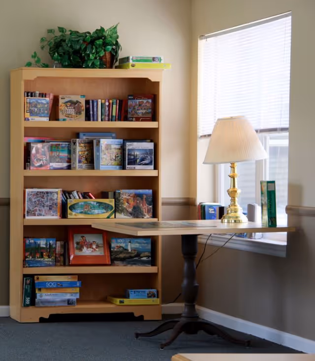 A cozy corner of a room featuring a wooden bookshelf filled with puzzles and books, a small table with a puzzle in progress, and a brass table lamp with a white lampshade next to a window with white blinds.