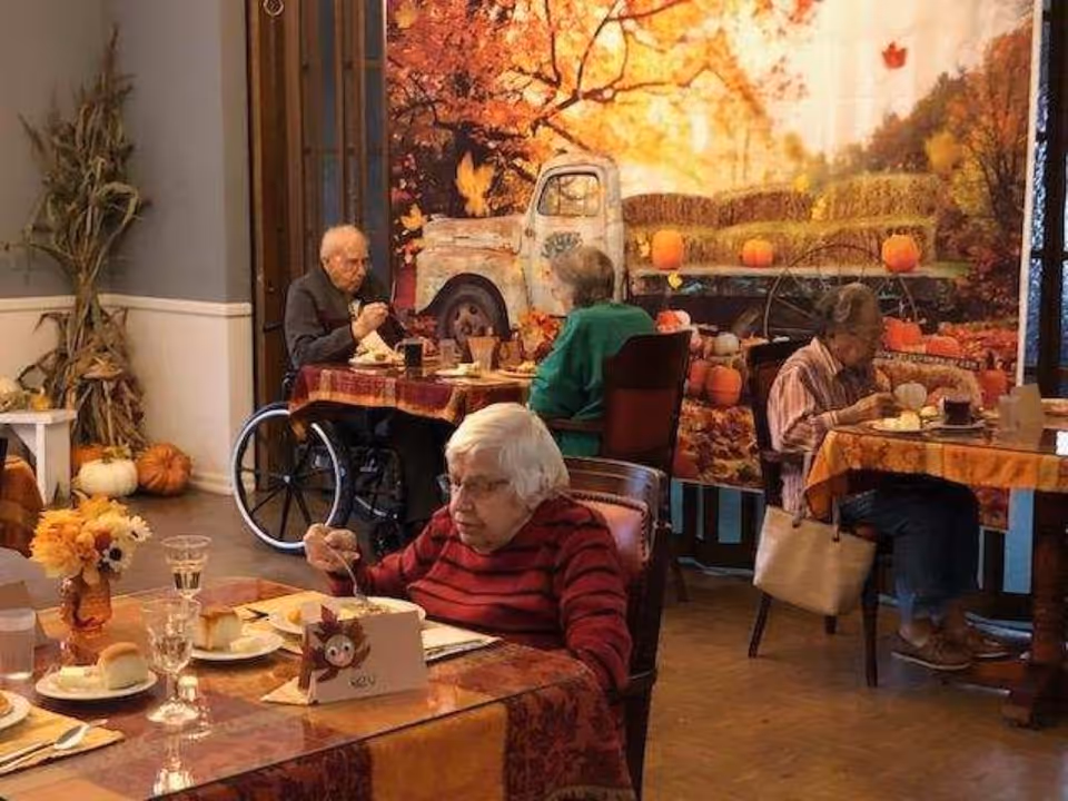 Four elderly adults seated at separate tables in a dining room decorated with autumn-themed tablecloths and a large fall backdrop featuring pumpkins and an old truck. One person is in a wheelchair, and the room has warm, cozy lighting with fall decorations including pumpkins and dried cornstalks.
