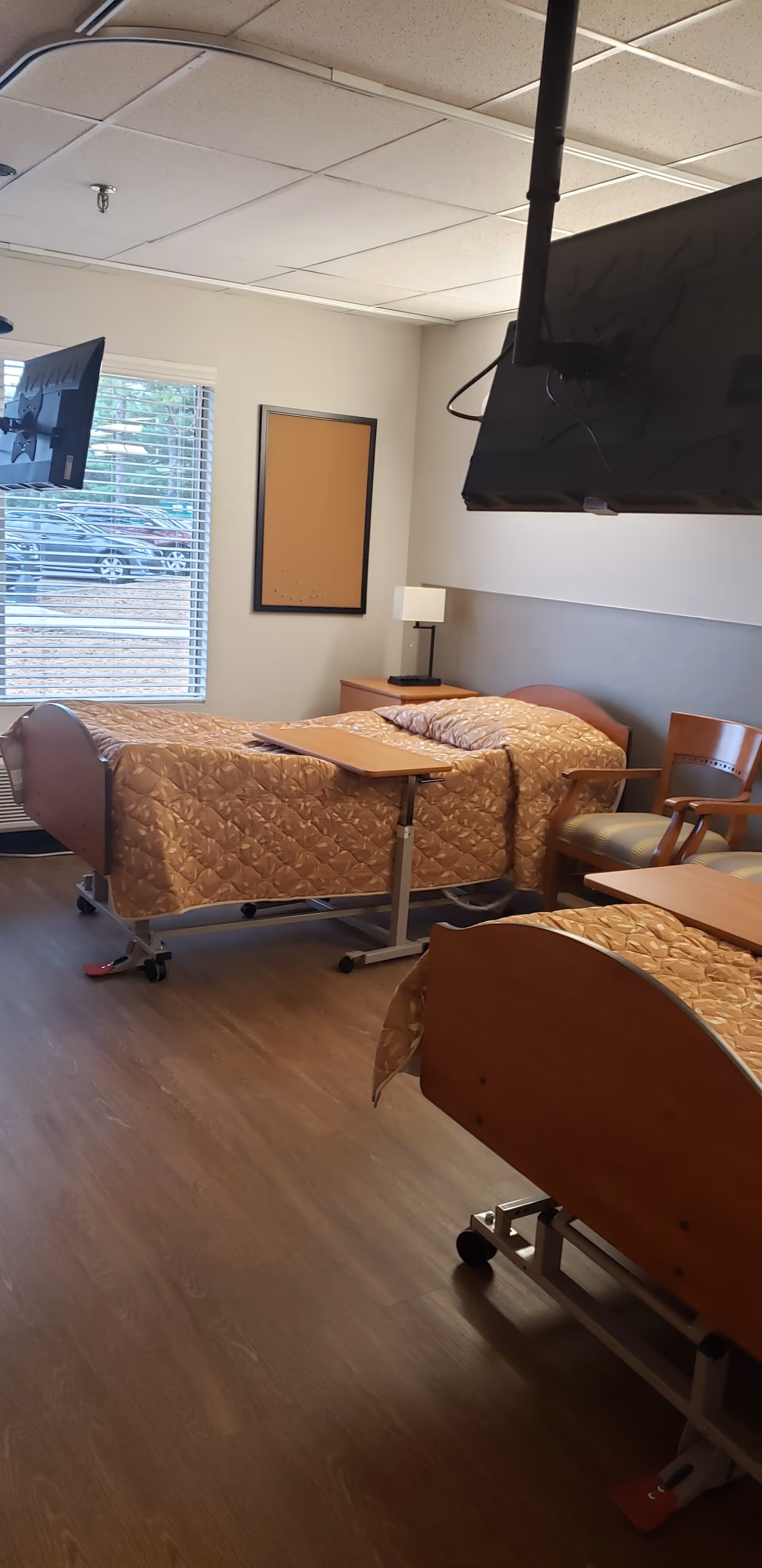 A nursing facility room with two hospital-style beds covered in matching beige patterned bedspreads. Each bed has an adjustable overbed table. There is a wooden chair next to one bed and a small nightstand with a lamp between the beds. Two wall-mounted televisions are visible, one near the window with blinds and another hanging from the ceiling. The room has wood-look flooring and a white ceiling with fluorescent lighting.