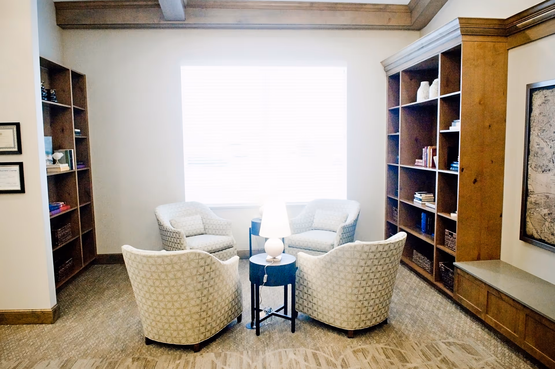 A cozy sitting area with four patterned armchairs arranged around a small round table with a lamp. The room has large wooden bookshelves filled with books and decorative items, a large window with blinds letting in natural light, and a framed map on the wall.