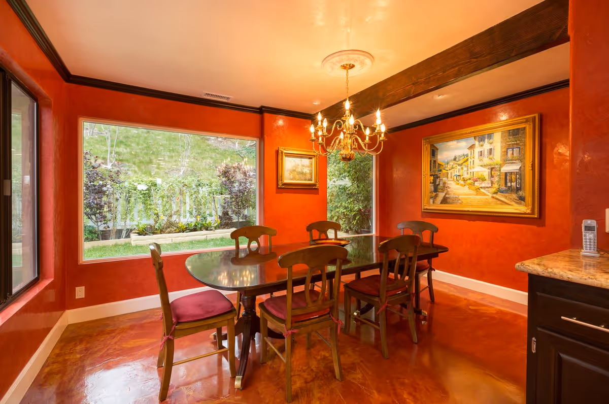 A dining room with red textured walls and a polished reddish-brown floor. There is a dark wooden dining table with six matching chairs that have red cushions. A gold chandelier with lit bulbs hangs above the table. Two large windows provide a view of green plants and a grassy hill outside. The walls are decorated with two framed paintings depicting outdoor scenes. A granite countertop with a cordless phone is visible on the right side.