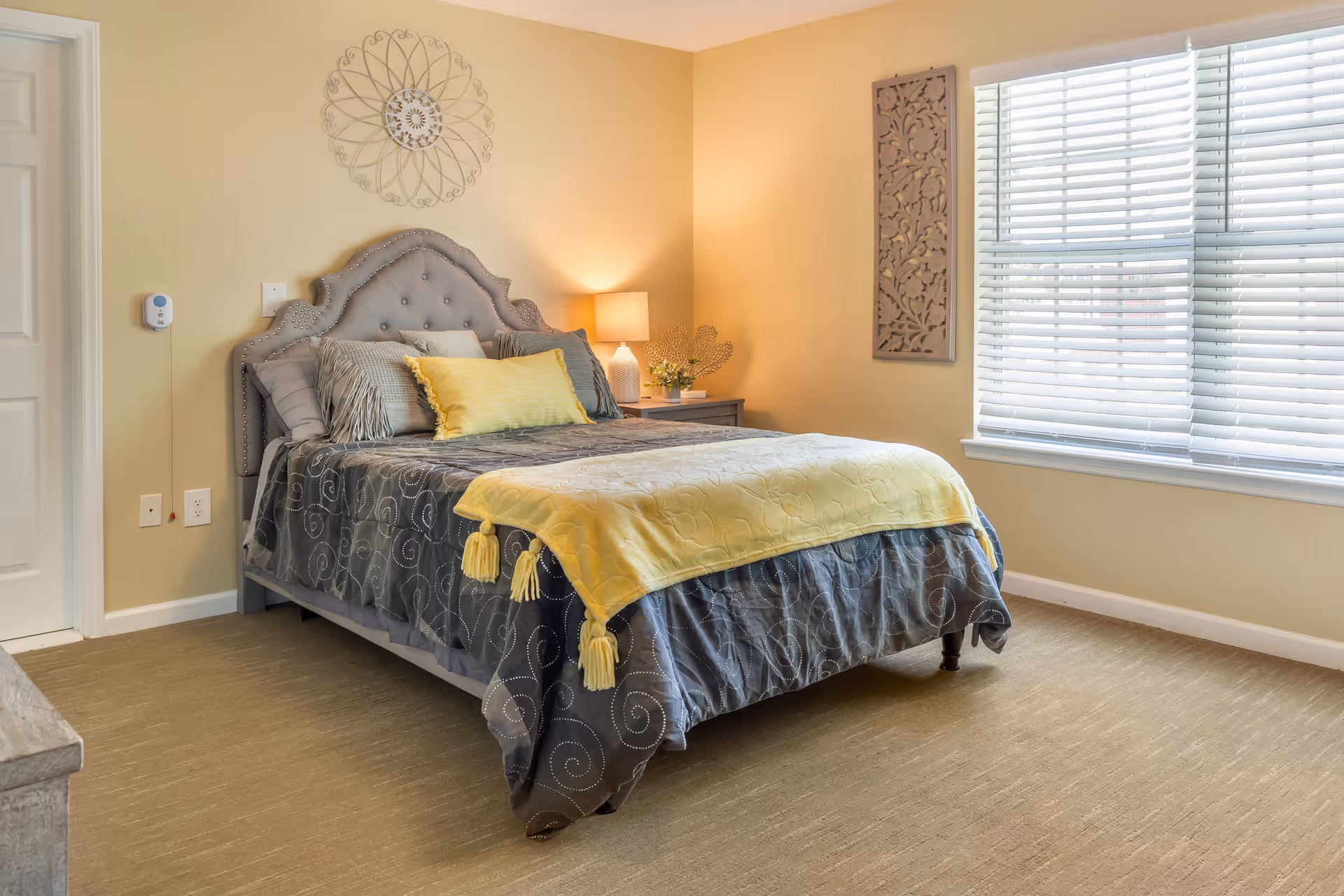 A neatly made bed with a tufted gray headboard in a bedroom. The bed has gray and yellow bedding with decorative pillows. A nightstand with a lamp and small plant is beside the bed. The room has beige walls, a window with white blinds, and a decorative wall hanging above the bed and on the adjacent wall.
