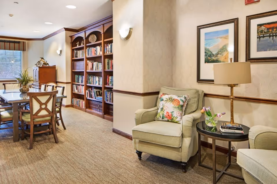 A cozy common area in a senior living facility featuring a comfortable armchair with a floral pillow, a small round side table with a lamp, books, and a vase with flowers. In the background, there is a wooden dining table with chairs and a large built-in bookshelf filled with books. The walls are decorated with framed artwork and the room has carpeted flooring.
