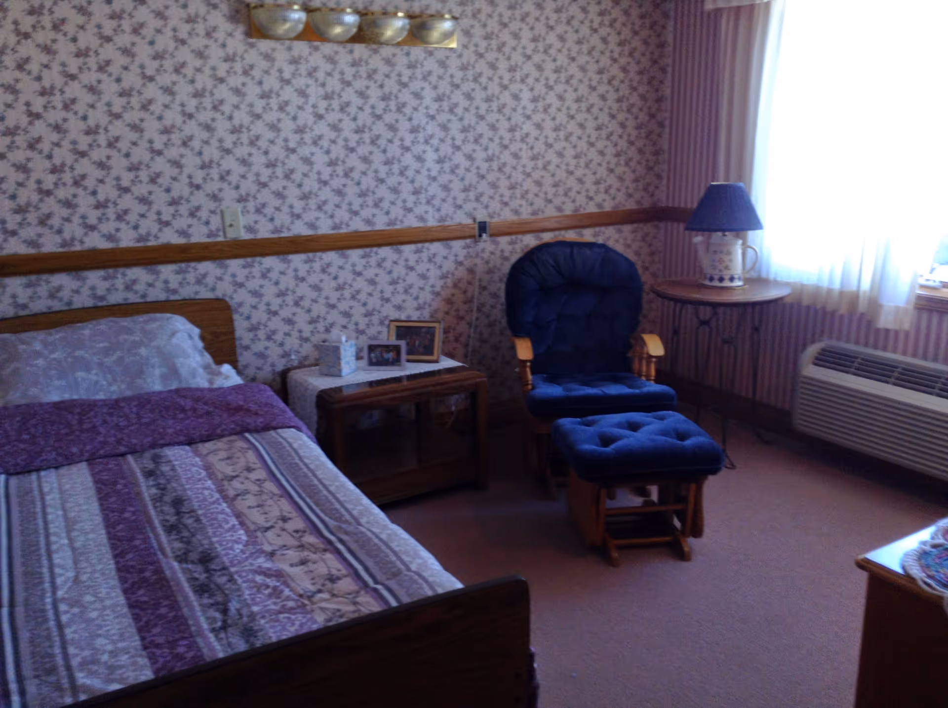A cozy senior living bedroom with a single bed covered in a purple and beige patterned quilt, a wooden nightstand with framed photos and a tissue box, a cushioned rocking chair with matching footstool, a round side table with a lamp, and a window with sheer curtains letting in natural light.