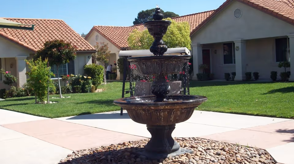 Outdoor courtyard area of a retirement center featuring a three-tiered water fountain surrounded by smooth stones, with green lawns, small trees, and single-story buildings with red tile roofs in the background.