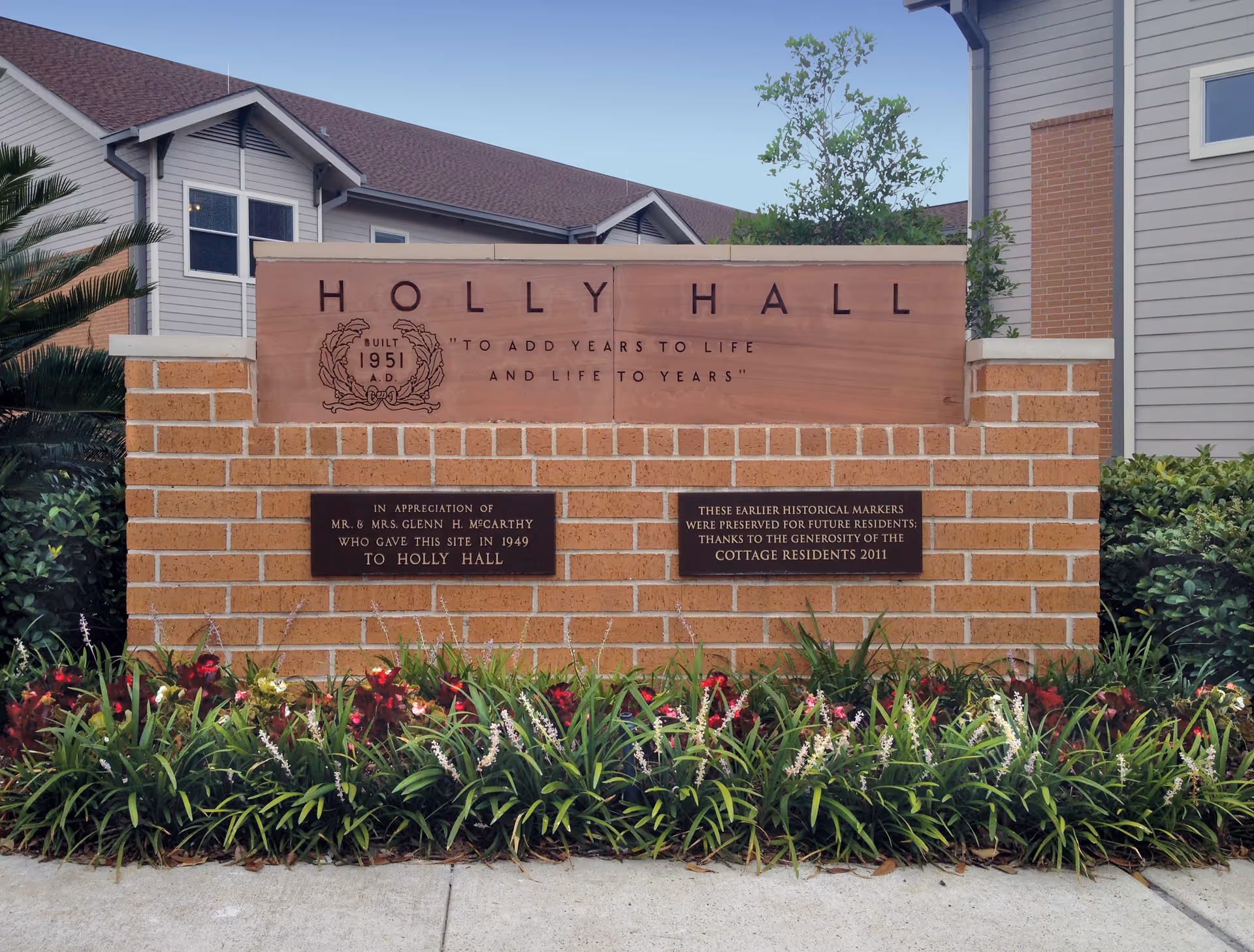 A brick sign for Holly Hall with the inscription 'To add years to life and life to years' and plaques honoring Mr. & Mrs. Glenn H. McCarthy and the generosity of cottage residents. The sign is surrounded by flowers and greenery with a building in the background.