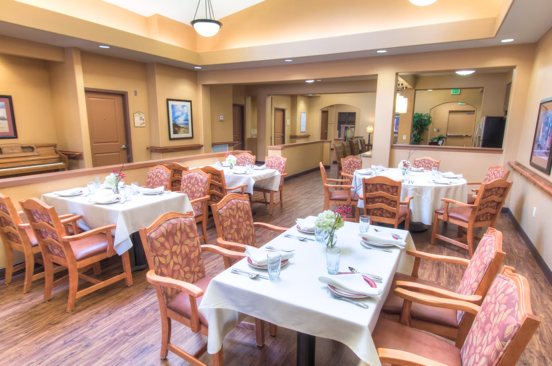 A dining room in a senior living facility with several tables covered in white tablecloths, each set with plates, glasses, napkins, and silverware. The chairs have wooden frames with cushioned seats and backs featuring a floral pattern. The room has warm beige walls, wood flooring, framed artwork, and a piano in the corner.