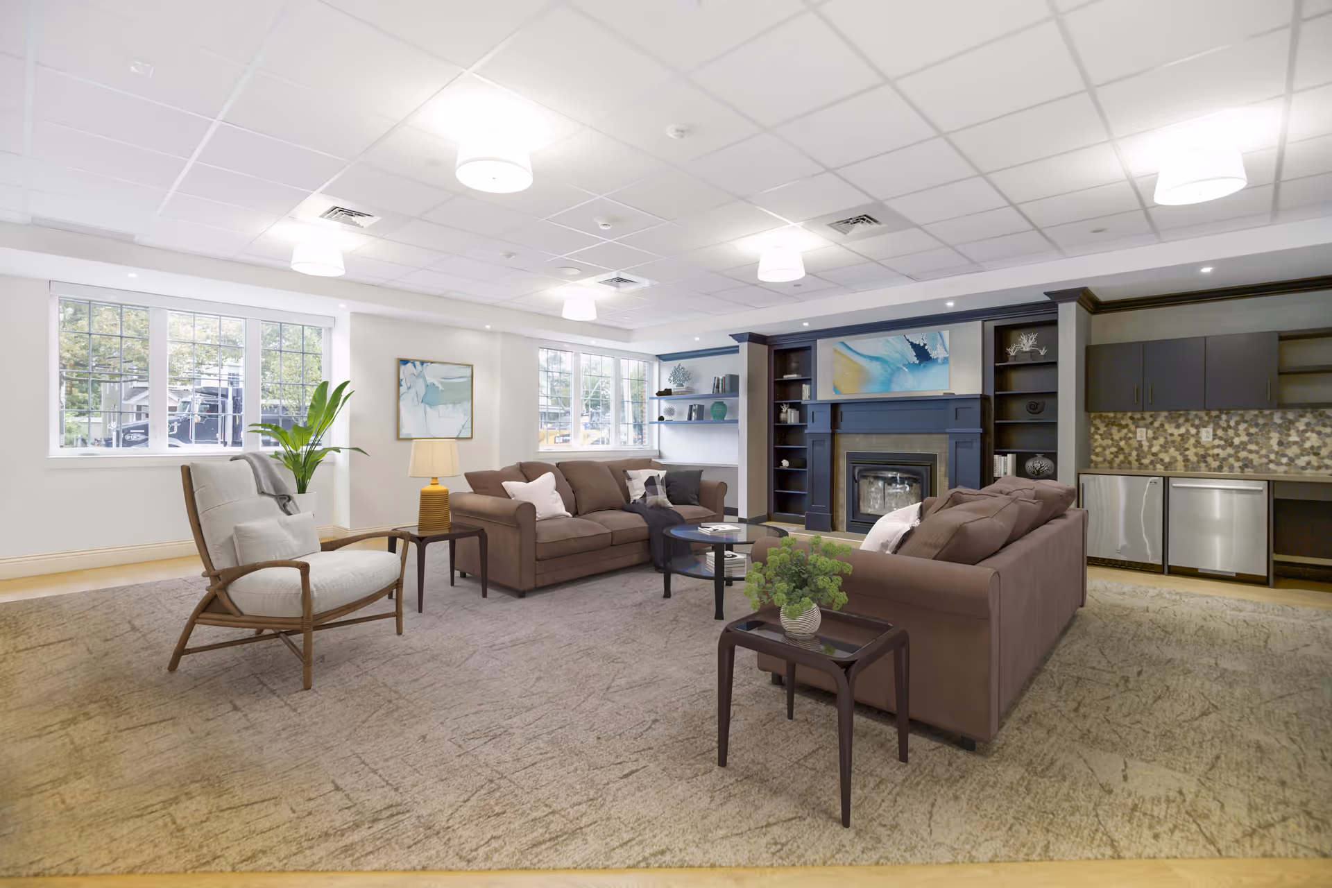 Spacious, well-lit communal living room with brown sofas and an armchair arranged around a fireplace and a small kitchenette along the back wall.