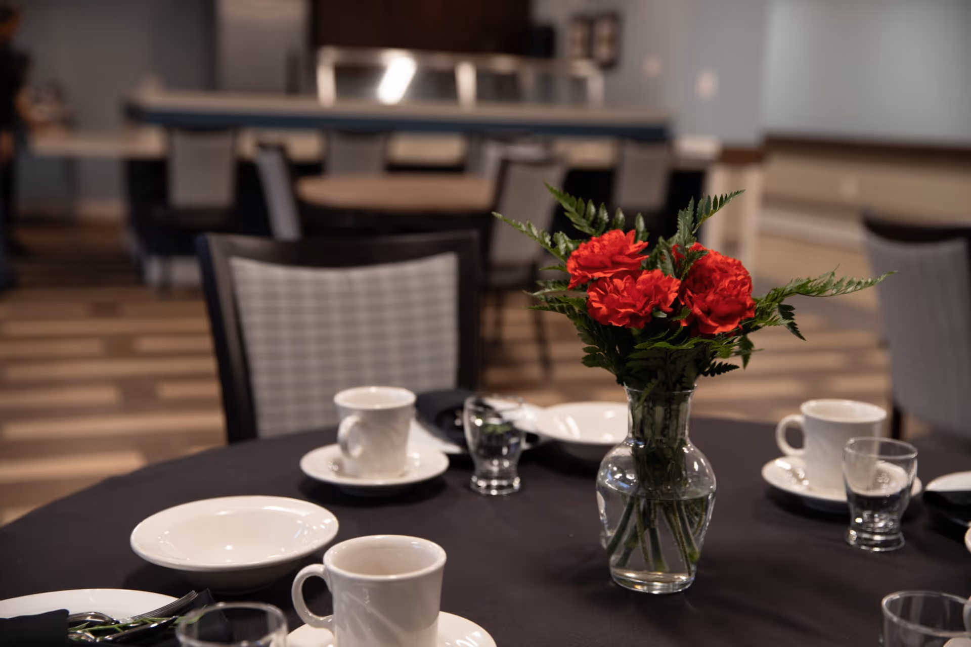 A dining table set with white dishes and a vase of red flowers in a communal dining room.