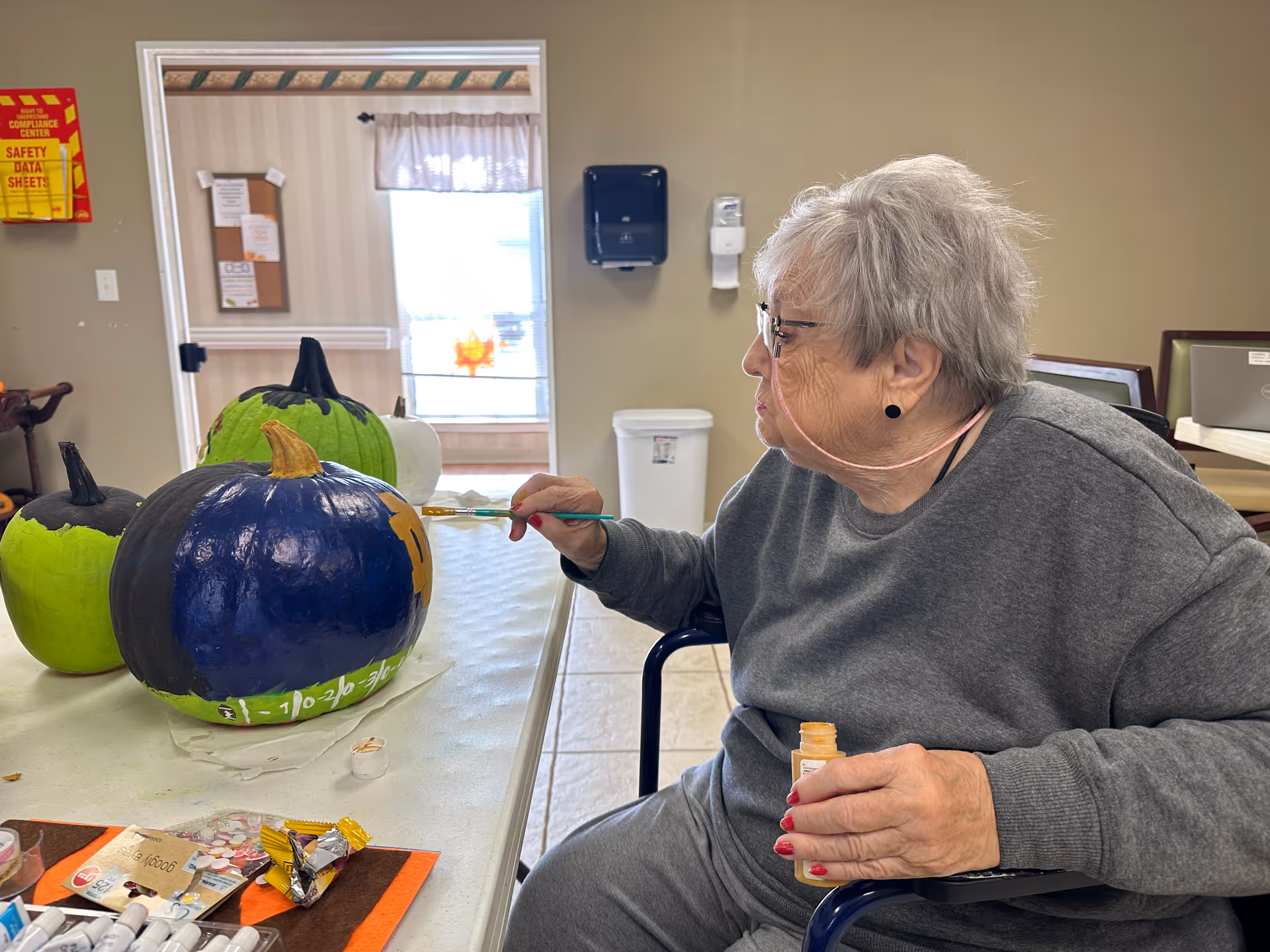 An elderly woman with gray hair and glasses is sitting in a wheelchair at a table, painting a pumpkin with a paintbrush. There are three pumpkins on the table, two painted green and one painted dark blue with some orange paint. The room has beige walls and a window with a curtain in the background.