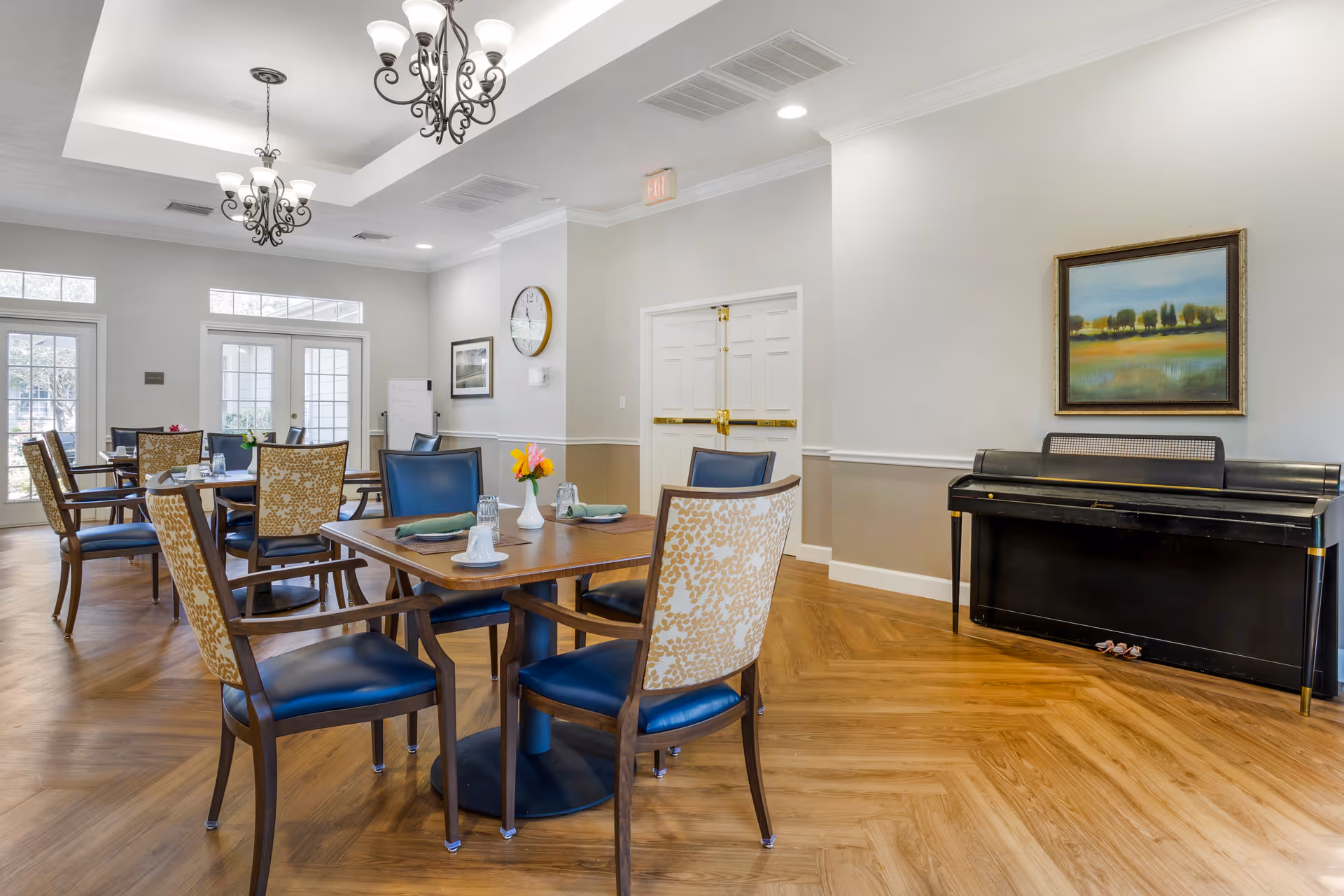 A bright dining room in a senior living facility with wooden floors, multiple tables set with cups and napkins, and chairs with blue seats and patterned backs. The room features elegant chandeliers, large windows and glass doors letting in natural light, a black piano against the wall, and a framed landscape painting above the piano.