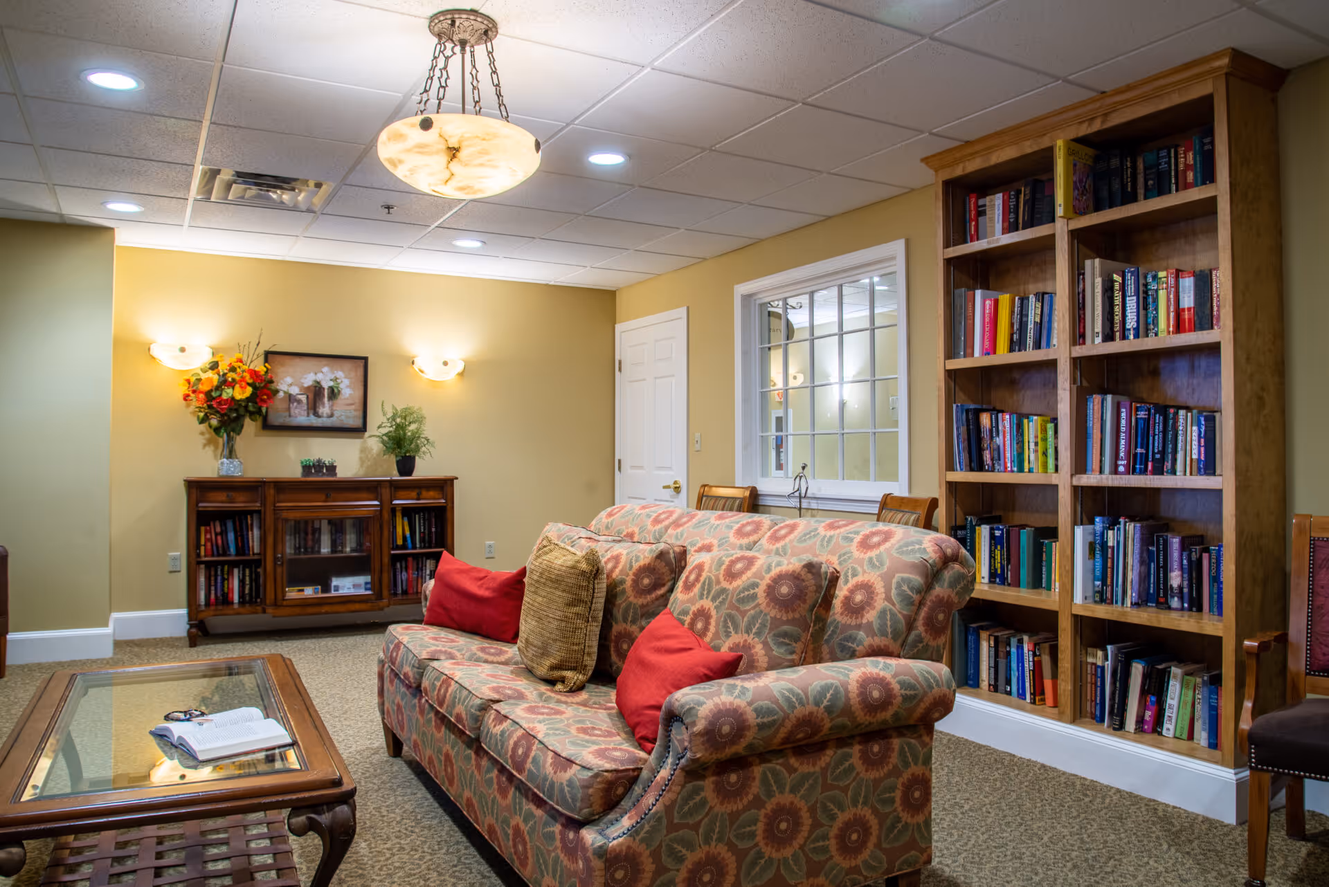 A cozy living room area with a patterned sofa adorned with red and beige cushions, a wooden coffee table with an open book, a tall wooden bookshelf filled with books, a wooden cabinet with decorative plants and a framed picture on the wall, warm yellow walls, and ceiling lights including a hanging light fixture.