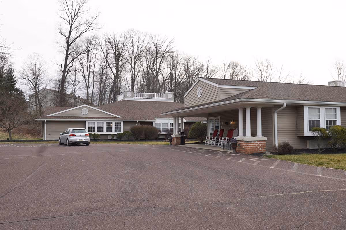 Exterior view of Celebration Villa of Exeter showing a single-story building with a covered entrance supported by white columns. There are several red rocking chairs on the porch. A silver car is parked in the large paved parking area in front of the building. Leafless trees and a cloudy sky are visible in the background.