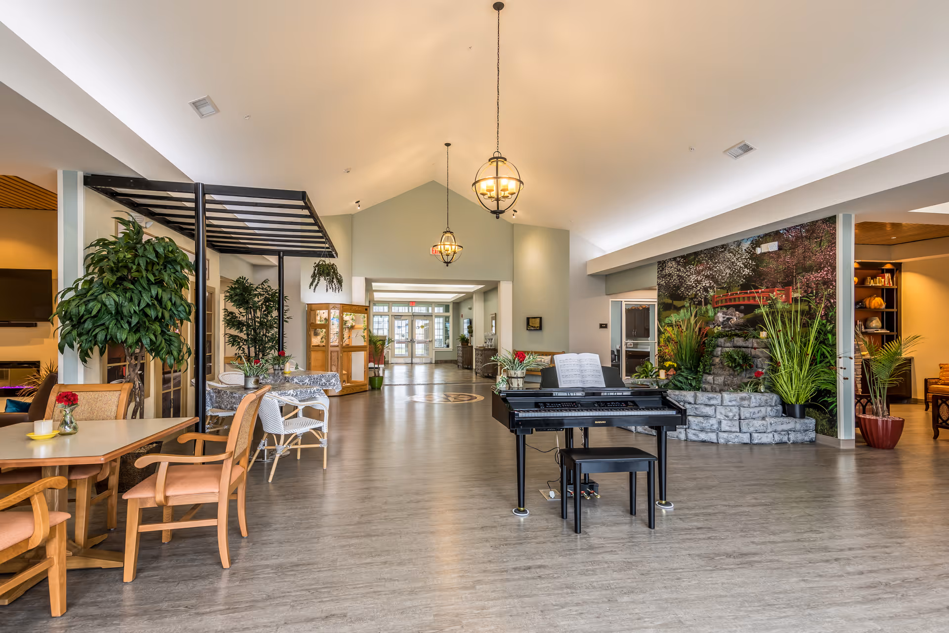 Spacious senior living common area with a black piano at center, seating tables, plants, and decorative hanging lights.