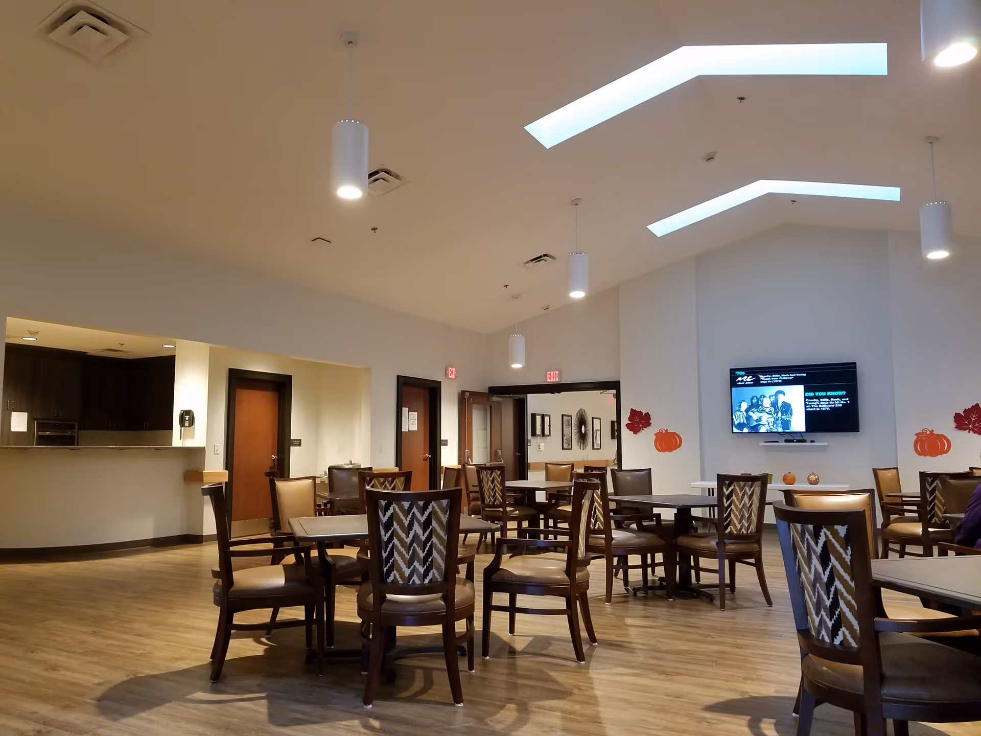 A spacious dining room with multiple wooden tables and chairs arranged neatly on a wooden floor. The ceiling has modern pendant lights and skylights. There is a wall-mounted TV displaying a program, and seasonal decorations such as pumpkins and leaves are on the walls. Doors and a pass-through window to a kitchen area are visible in the background.