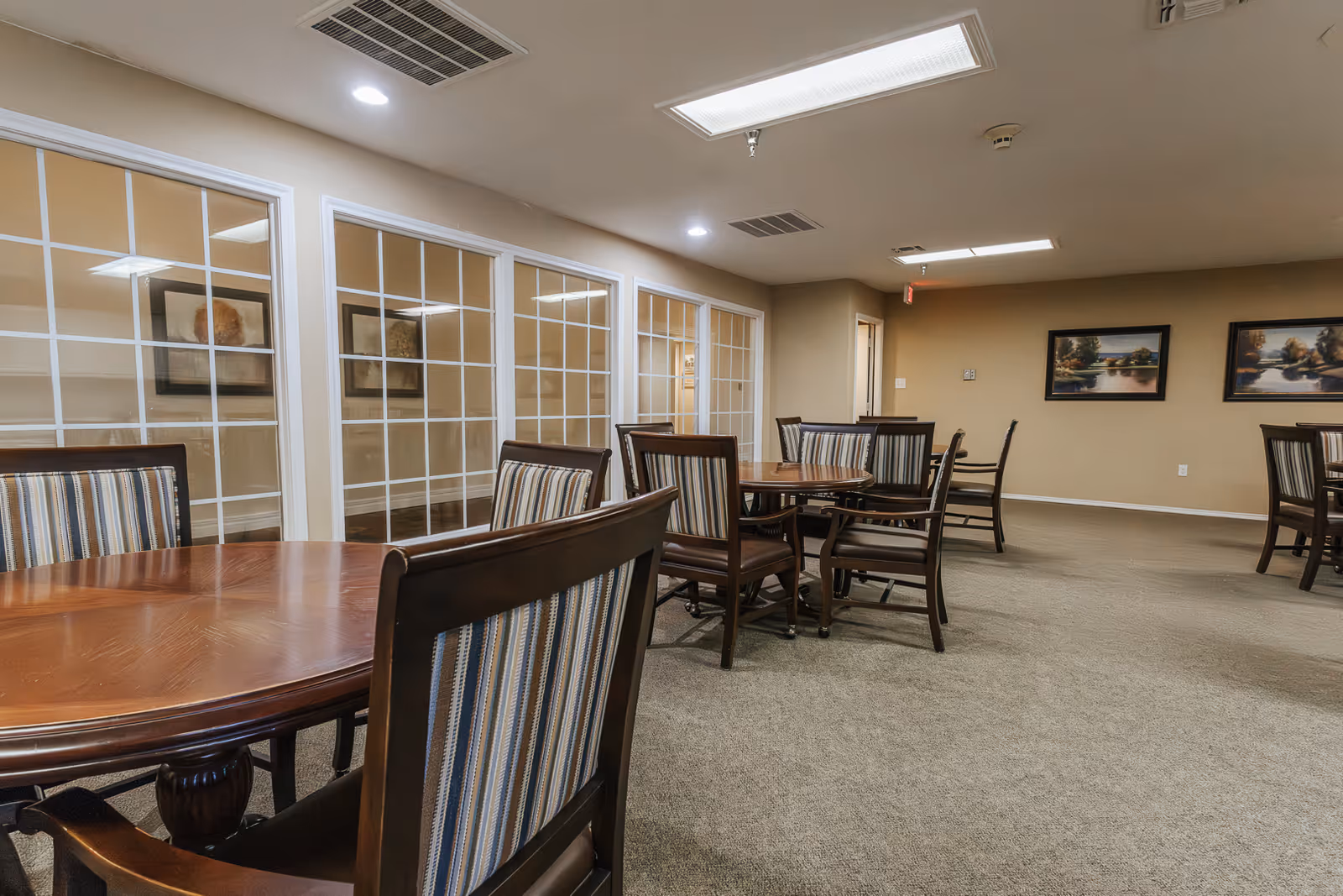Interior view of a senior living facility dining room with several round wooden tables and striped cushioned chairs. The room has beige walls, carpeted floor, framed artwork on the walls, and large windows with white grid frames on one side.