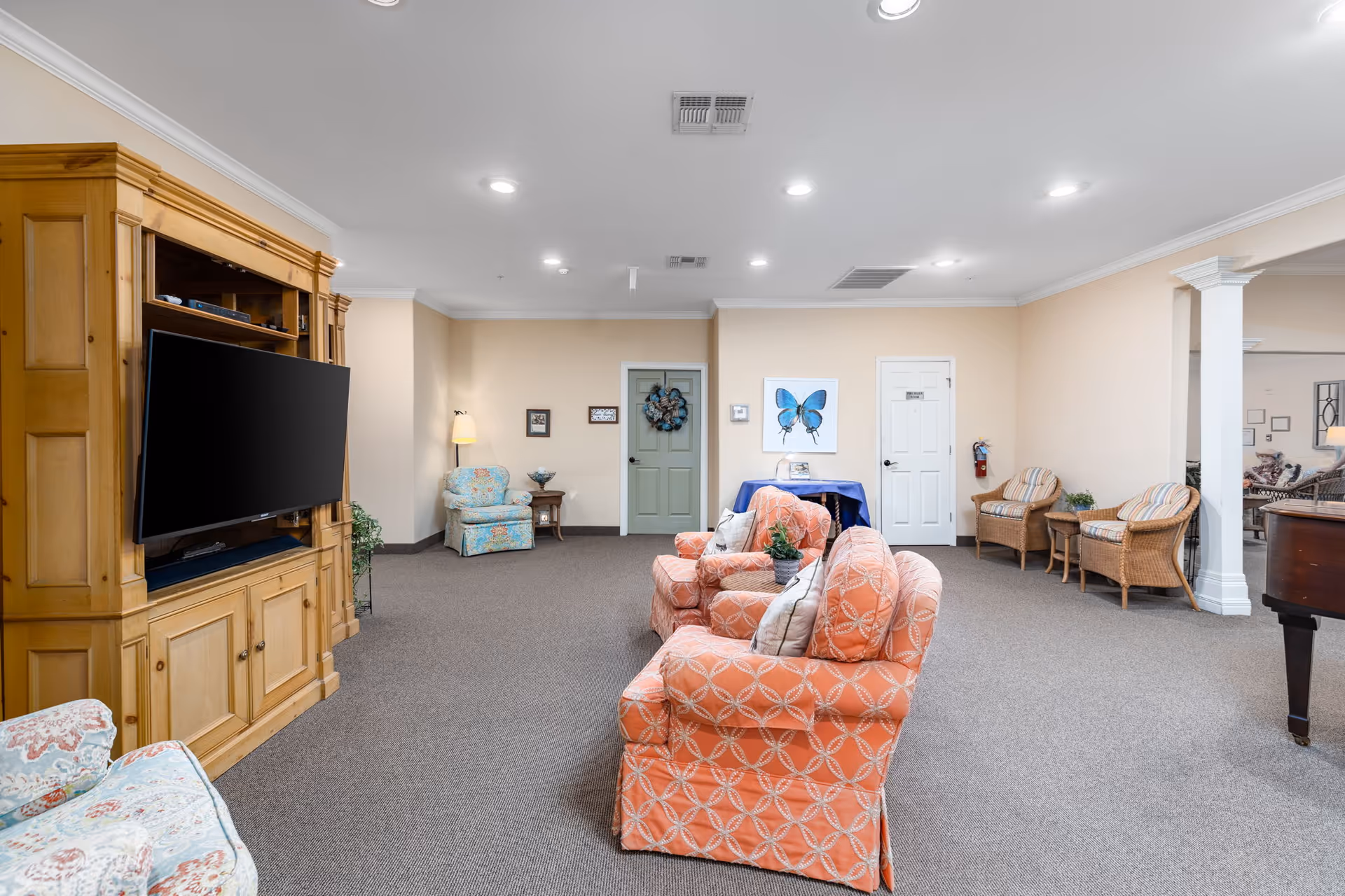 A spacious living room area with beige walls and carpeted floor. The room features a large wooden entertainment center with a flat-screen TV, two coral patterned armchairs with a small plant on the armrest, a floral armchair near a floor lamp and side table, and two wicker chairs with striped cushions. There is a table covered with a blue cloth against the wall, decorated with a framed blue butterfly artwork. Two doors are visible on the far wall, one painted green and the other white. The ceiling has multiple recessed lights and air vents.
