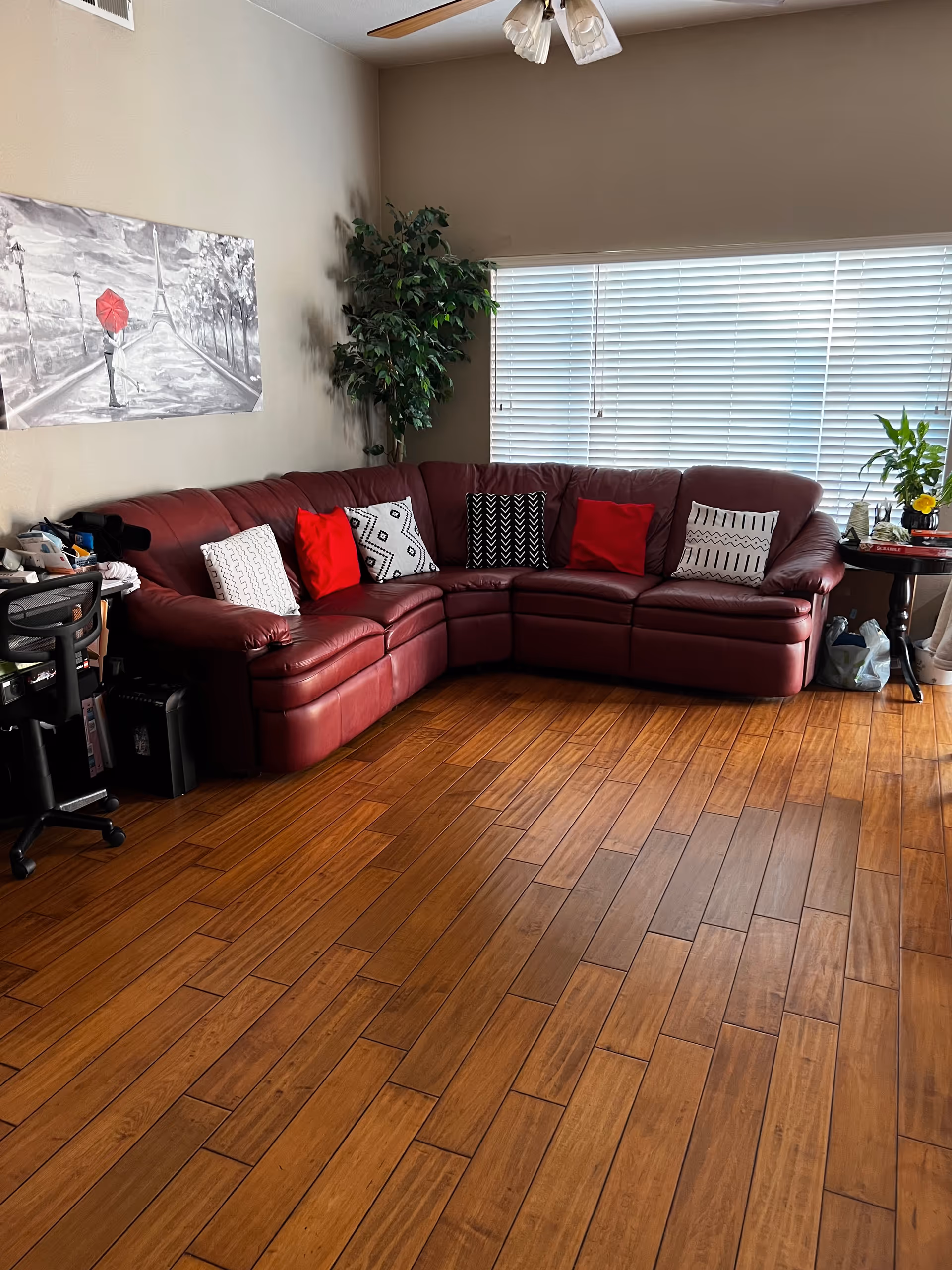 A cozy living room with a large red leather sectional sofa adorned with patterned and solid red pillows. There is a tall green plant in the corner, a ceiling fan with lights above, and a large window with white blinds letting in natural light. A black office chair and desk are partially visible on the left side, and a small round table with plants and books is on the right side. The floor is wooden with a warm finish.