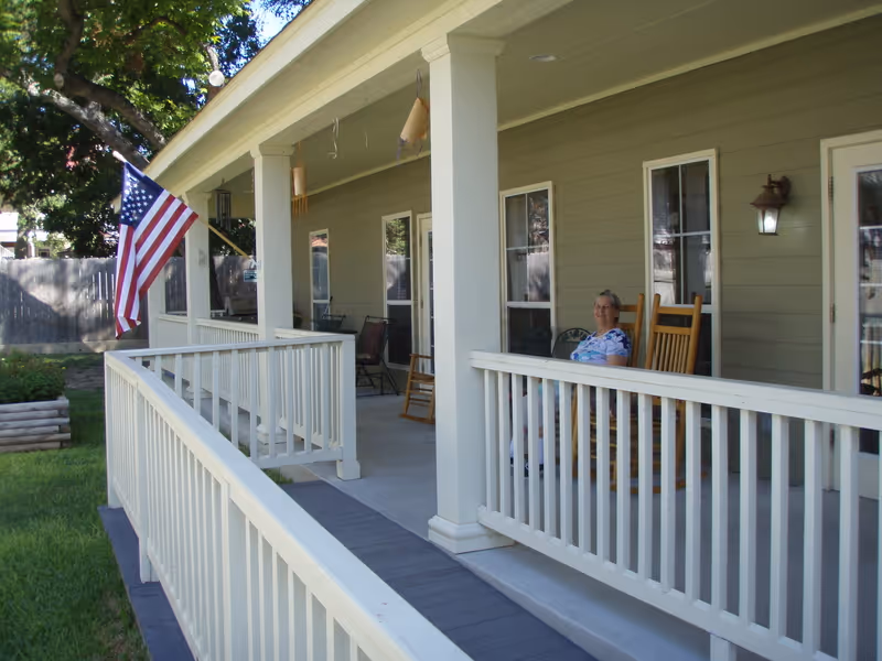 A covered porch area with white railings and columns. An elderly woman is sitting on a wooden rocking chair on the porch. There is an American flag hanging from one of the columns. The porch is attached to a green building with multiple windows and a door. The area around the porch has green grass and a tree providing shade.