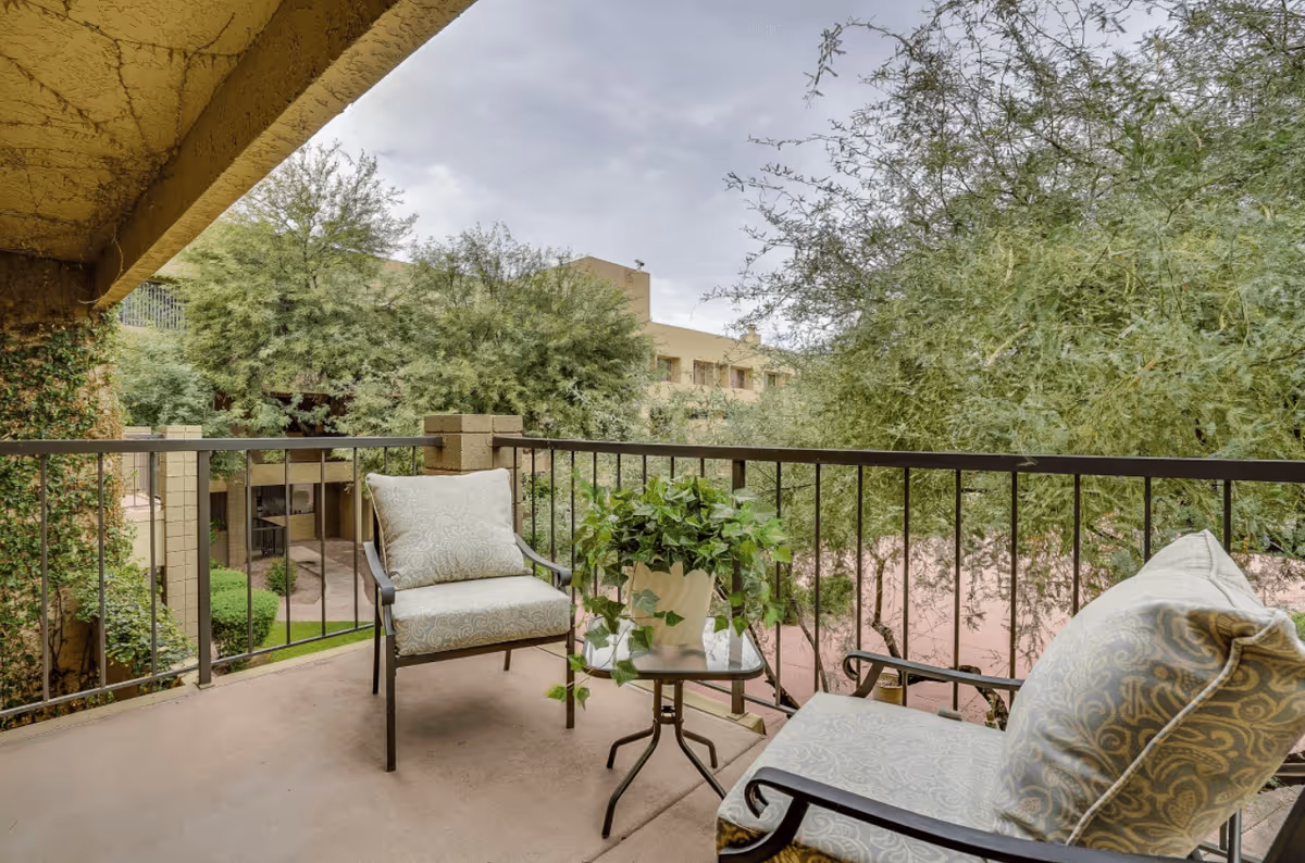 A covered outdoor balcony with two cushioned chairs and a small glass-top table holding a potted green plant. The balcony overlooks trees and a beige building under a cloudy sky.