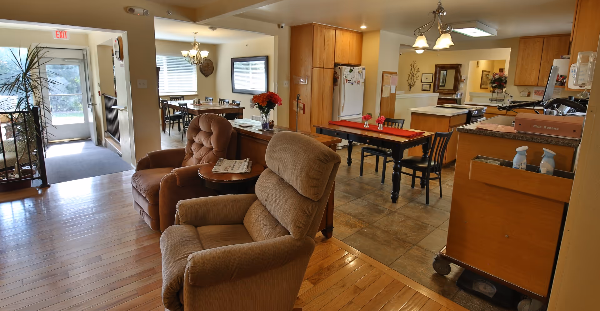 Interior view of a senior living facility showing a common area with two comfortable armchairs, a small round table with a newspaper, a dining table with chairs, and a kitchen area with wooden cabinets and appliances. The space is well-lit with natural light coming through windows and a glass door in the background.