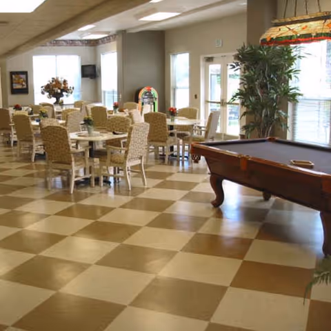 A spacious room with checkered tile flooring featuring several tables and chairs arranged for dining or socializing. A pool table is positioned on the right side near large windows with blinds, and a tall potted plant is placed beside the windows. The room is well-lit with natural light coming through the windows and has a cozy, inviting atmosphere.