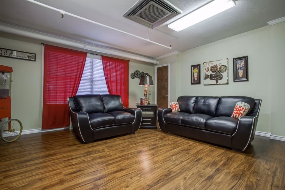 A living room with two black leather sofas, one loveseat and one three-seater, each with decorative pillows. There is a small black side table with a lamp between the sofas. The room has wooden flooring, light green walls, and a window with red curtains. Wall decorations include framed pictures and a vintage movie-themed sign. A popcorn machine is partially visible on the left side.