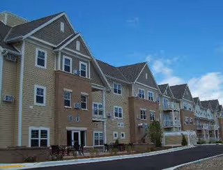 Exterior front of a multi-story beige and brick senior living building with balconies, many windows, and a paved driveway.