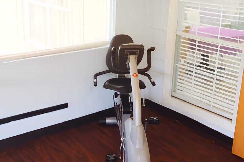 A stationary exercise bike with a black seat and backrest positioned in a corner of a room with wooden flooring and white walls. There is a window with white blinds on the right side and a large window with sheer curtains on the left side.