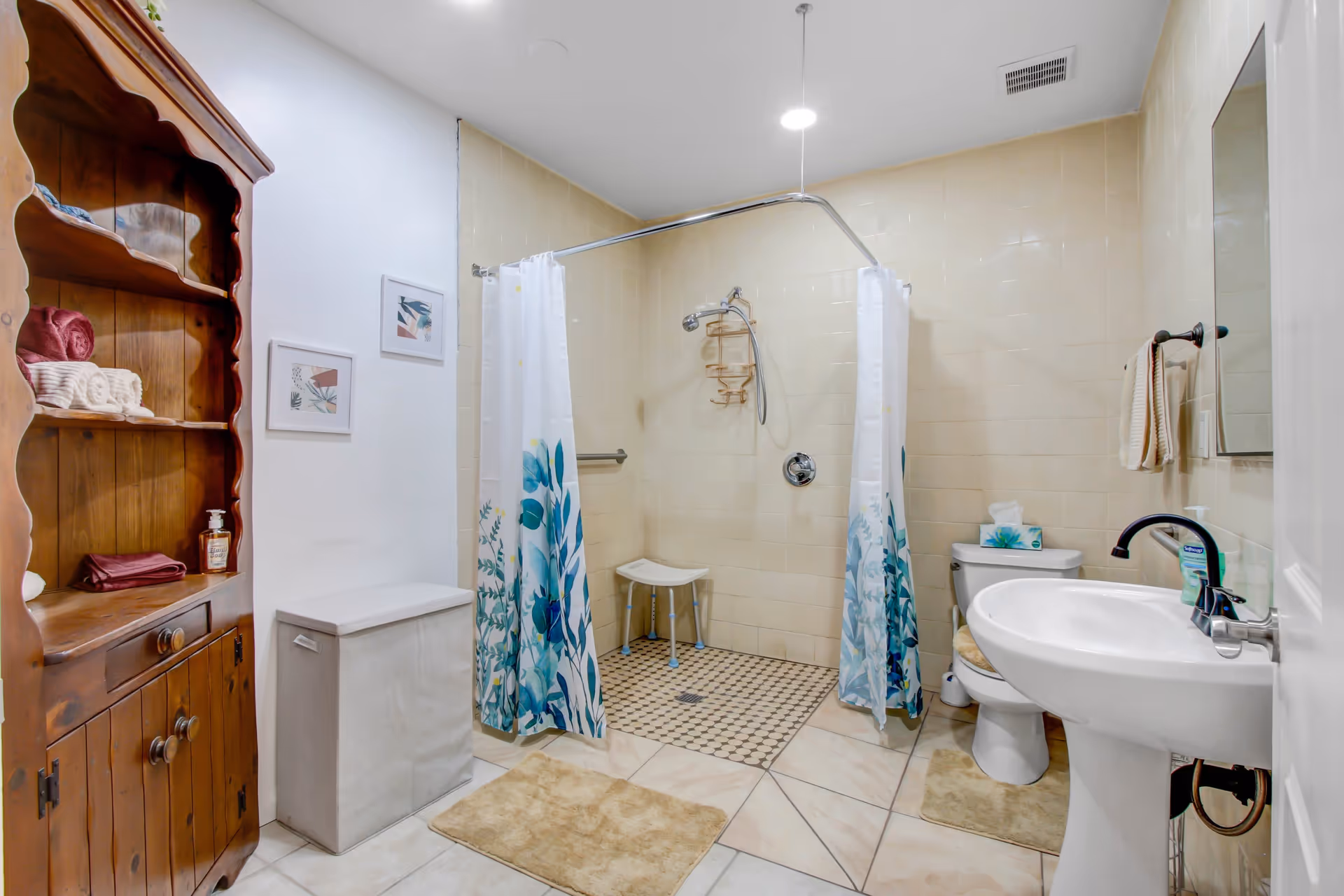 A clean and accessible bathroom with a walk-in shower featuring a white curtain with blue floral designs, a white shower stool, a toilet with a tissue box on top, a white pedestal sink with a black faucet, a wooden cabinet with towels and soap, two framed pictures on the wall, and beige tiled floors and walls.