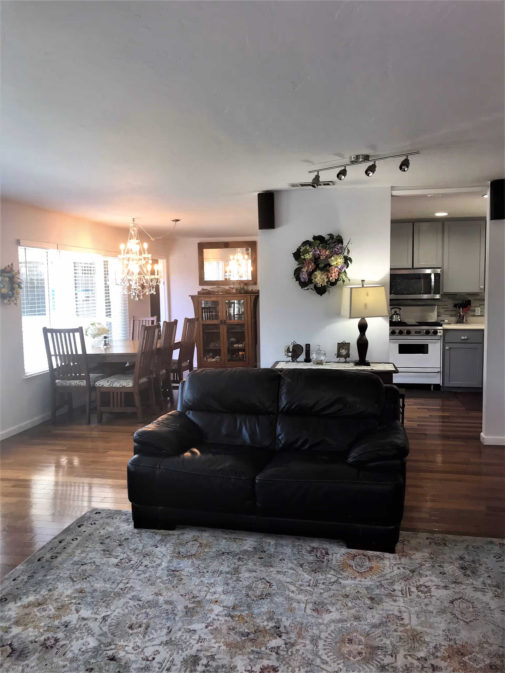 Interior view of a living space in Roses Assisted Living featuring a black leather loveseat on a patterned area rug in the foreground. Behind it, there is a dining area with a wooden table and chairs, a chandelier above the table, and a wooden cabinet with glass doors. To the right, a partial view of a kitchen with gray cabinets, a stove, and a microwave is visible. The walls are decorated with floral wreaths and a table lamp is placed on a console table against a white wall.