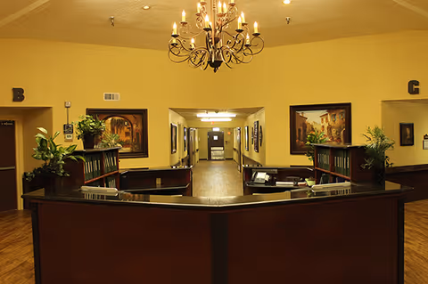 Interior view of a nursing and rehab facility reception area with a dark wood front desk, potted plants, framed artwork on yellow walls, a chandelier hanging from the ceiling, and a hallway leading to other rooms.