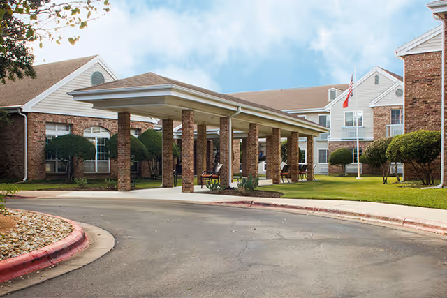 Entrance area of a senior living facility with a covered drop-off zone supported by brick columns, surrounded by well-maintained landscaping and brick buildings under a partly cloudy sky.